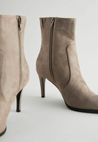 Botine din suede de culoare taupe, cu vârf ascuțit, fermoare pe lateral și tocuri stiletto de 10 cm. Textură netedă cu detalii de cusături vizibile.