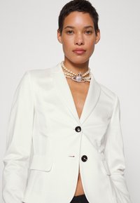 White tailored blazer with peaked lapels, featuring two black buttons, side pockets, and a smooth texture. Styled with a pearl choker.