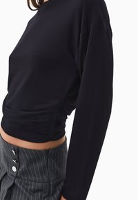 Kvinna i en figurnära svart långärmad topp med rynkad sidodetalj och högmidjade grå rutiga byxor med knappar och dragkedja.