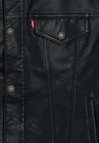 Giacca in pelle nera con bottoni a pressione, superficie testurizzata e una tasca sul petto ben visibile con un'etichetta rossa Levi's e dettagli di cucitura verticale.