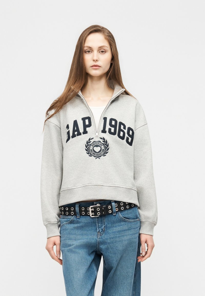 Jeune femme aux longs cheveux portant un sweat-shirt gris clair GAP 1969 à fermeture éclair quartée et un jean bleu avec une ceinture noire à œillets.