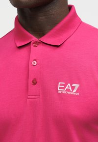 Άτομο με σκουρόχρωμο δέρμα που φοράει φωτεινό ροζ μπλουζάκι πόλο EA7 Emporio Armani με τρία κουμπιά και γιακά, ορατό από το λαιμό μέχρι το μέσο του στήθους.