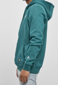 Sudadera con capucha color azul verdoso, con bolsillo frontal, capucha con cordón y logo de estrella blanca en la manga. Mezcla de algodón con una textura suave.