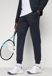 Tumši zili skrējēja stila bikses ar sānu kabatām, ribota stila potīšu apmali un fakturētu dizainu. Kombinētas ar baltām sporta kurpēm un tenisa raketi.