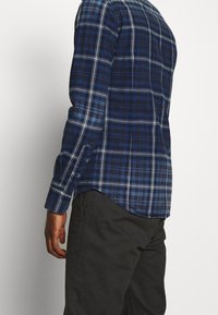 Uomo che indossa una camicia a maniche lunghe a quadri blu e nera e pantaloni scuri, in piedi di lato contro uno sfondo chiaro e semplice.