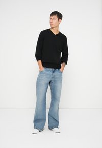 Svart V-hals genser, avslappet passform, kombinert med lyseblå vide jeans. Modell iført hvite sneakers, hendene i lommene, nøytral bakgrunn.