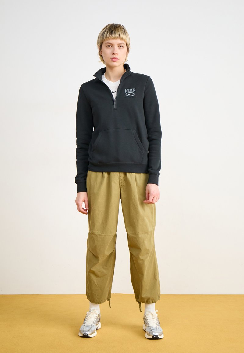 Sudadera negra con cremallera y bolsillo frontal, combinada con pantalones sueltos de color verde oliva claro y zapatillas deportivas grises.