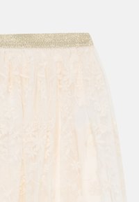 Spitzenrock mit einer durchscheinenden beigen Textur und einem glitzernden goldenen Taillenbund, der mit floralem Muster über den Stoff verziert ist.