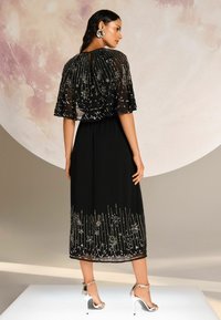 Juoda suknelė su permatomu, siuvinėjimu dekoruotu apsiaustu, puoštu sidabriniais gėlės raštais. Kelių ilgumo sijonas su karoliukais apatinėje dalyje. Sidabrinės aukštakulnės šlepetės.