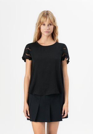 Jeune femme aux cheveux blonds portant un haut noir à manches courtes avec des détails en broderie et une jupe plissée noire, se tenant devant un fond blanc.