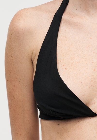 Κοντινό πλάνο ενός ατόμου που φοράει μαύρο μπικίνι με halter, δείχνοντας τον ώμο, το πάνω μέρος του στήθους και μέρος του κορμού σε απλό φόντο.