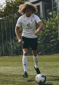 Maglietta da calcio bianca con accenti marroni, pantaloni corti neri e calze bianche. Il giocatore controlla un pallone da soccer decorato sull'erba, con una rete dietro.