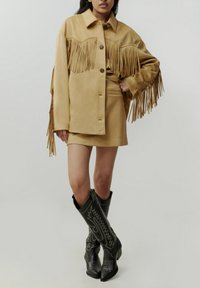 Beige Fransen-Jacke aus Kunstleder mit auffälligen Knopfverschlüssen, getragen über einem passenden Rock. Schwarze Cowboy-Stiefel mit dekorativen Stickdetails.
