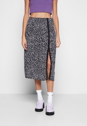 Vrouw draagt een zwarte midirok met witte abstracte patronen en een splits aan de voorkant over een paarse cropped top, witte sokken en lavendelkleurige loafers.