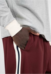 Sivá košeľa s dlhým rukávom a krémovými manžetami, skombinovaná s marhuľovými nohavicami s bočným pruhom v námorníckej a krémovej farbe. Ruka spočíva v vrecku.