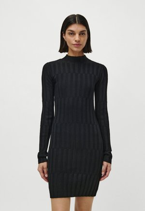 Woman with straight dark hair wears a fitted black ribbed knit dress with long sleeves and a high neckline, standing against a plain background.