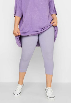 Personne portant des leggings courts violet clair, un haut violet surdimensionné et des baskets blanches, tenant l'ourlet du haut avec les deux mains.