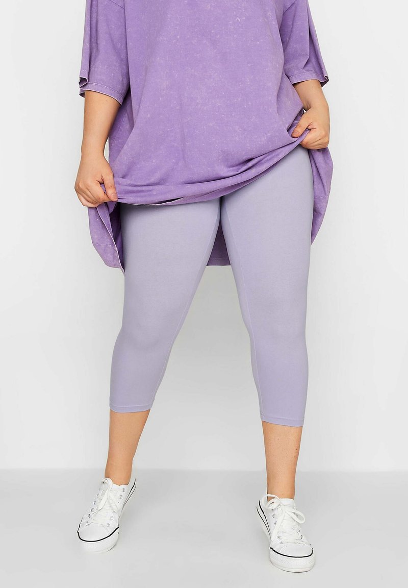 Personne portant des leggings courts violet clair, un haut violet surdimensionné et des baskets blanches, tenant l'ourlet du haut avec les deux mains.