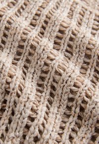 Strikket tekstil i lys beige med et tekstureret mønster, der indeholder tynde, skinnende tråde vævet ind for en subtil glans.