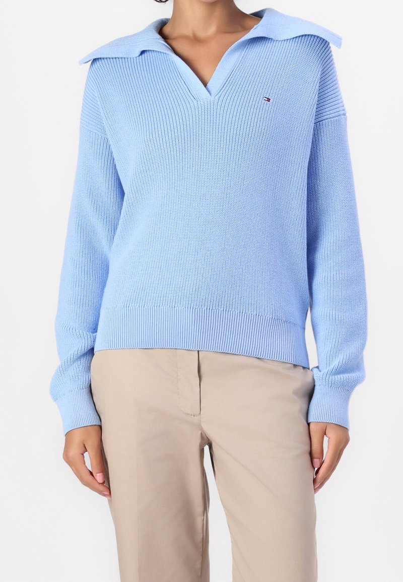 Femme portant un pull en tricot bleu clair avec un col large et un pantalon beige, debout devant un fond blanc uni.