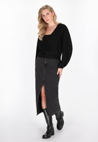 Frau mit blonden, gewellten Haaren, die einen schwarzen, flauschigen V-Ausschnitt-Pullover, einen taillenhohen schwarzen Midirock mit Schlitz vorne und schwarze Stiefel bis zu den Knien trägt.