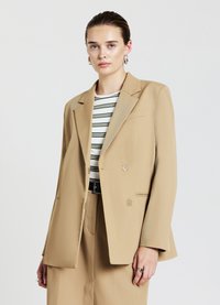 Blazer beige doppiopetto con rever a punta, tessuto testurizzato e due bottoni decorativi. Modello che indossa una maglietta a righe sotto.