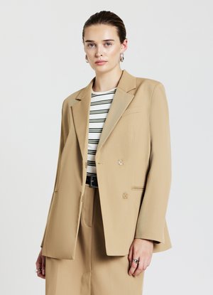 Beige Doppelreiher-Blazer mit spitzen Revers, strukturiertem Stoff und zwei dekorativen Knopfakzenten. Modell trägt ein gestreiftes Oberteil darunter.