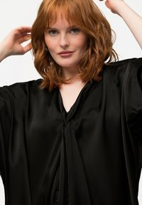 Blusa di raso nero con scollatura a V, maniche morbide e dettaglio a fiocco sul collo. Texture liscia con una finitura lucida.