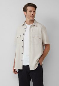 Korte mouwen, crèmekleurig shirt van lichtgewicht stof, voorzien van donkere knopen, twee borstzakken en een kraag.