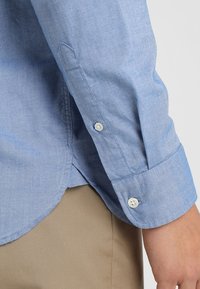 Poignets de chemise boutonnée à manches longues bleu clair sur un pantalon beige, montrant la couture latérale et les boutons sur la manche près du poignet.