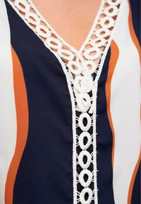 Blouse marine avec un décolleté en V, ornée de dentelle crochet blanche et de rayures verticales multicolores en orange et écru. Tissu doux.