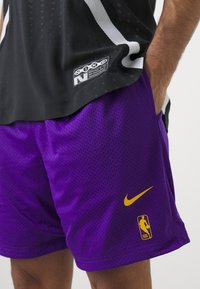 Črn mrežast dres z belimi akcenti; vijolične perforirane kratke hlače z rumenim Nike swoosh-om in NBA logotipom. Idealno za športno oblačenje.