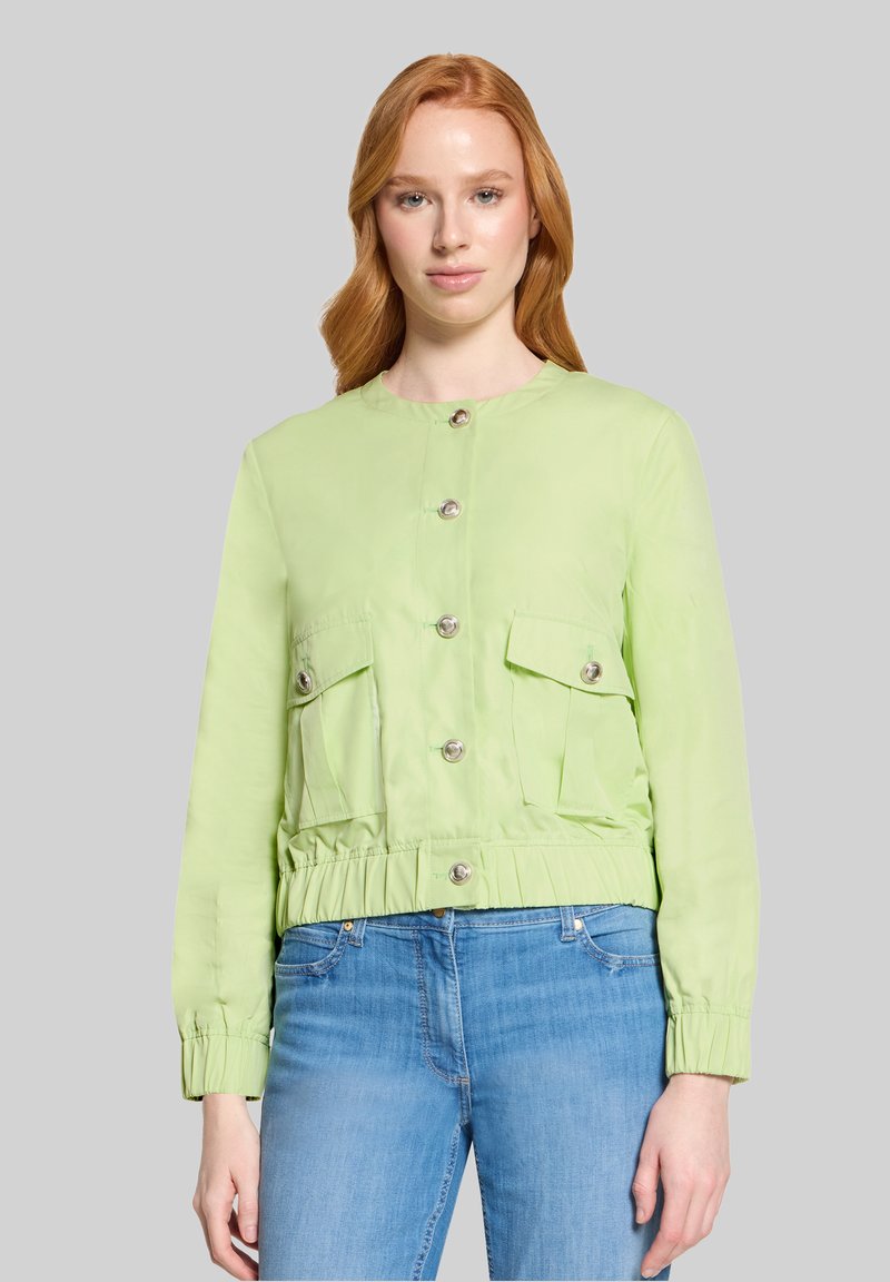 Femme aux longs cheveux roux portant une veste verte claire boutonnée avec deux poches avant et un jean bleu, debout devant un fond gris.