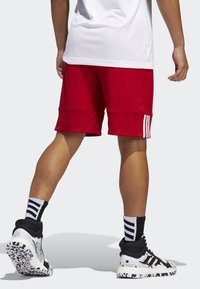 Shorts de sport rouges avec une texture lisse, ornés de trois rayures verticales blanches sur le côté gauche. Portés avec des baskets à motifs noir et blanc.