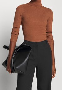 Pull à col roulé côtelé marron à manches longues associé à un pantalon noir taille haute. Un grand sac à main noir et doux est tenu dans une main.