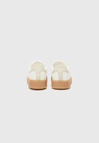 White sneakers with a textured cream suede panel, tan rubber soles, and embossed branding on the heel. Simple, low-profile design.