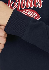 Sudadera de color azul marino con puños acanalados, que presenta un diseño de texto gráfico en rojo y blanco. Material suave y texturizado con una forma ajustada.