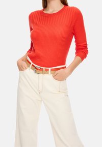 Pull rouge côtelé à col rond, associé à un pantalon large crème et une ceinture beige clair. Présente une silhouette ajustée et des manches retroussées.