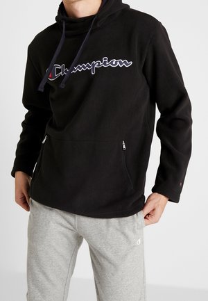 Hombre con sudadera negra Champion de forro polar con capucha, bolsillos frontales con cremallera y pantalones de chándal grises en un ambiente interior casual.