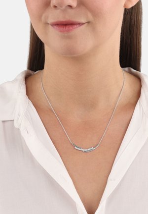 Collana d'argento con pendente a barra curvata decorata con piccoli pietre trasparenti, indossata da una donna con capelli castani lisci e una camicia bianca con colletto.