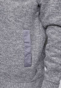 Gros plan sur un pull en tricot texturé gris avec une poche en tissu lisse verticale sur le côté et des bords et des poignets côtelés.