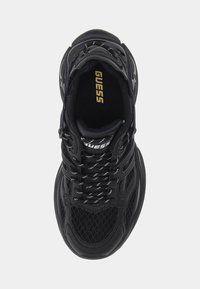 Musta urheilukenkä, jossa on verkkokangasta ja synteettisiä materiaaleja, pehmustettu kaulus ja valkoiset ompeleet. Pohjallisessa kultainen "GUESS"-logo.