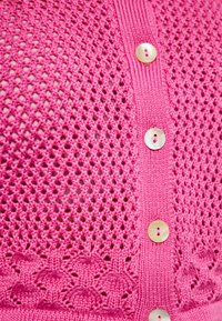 Ružová pletená kardigán s mriežkovaným vzorom. Obsahuje zapínanie na gombíky s okrúhlymi, svetlo sfarbenými gombíkmi a dekoratívnym pleteným lemom na spodnej časti.