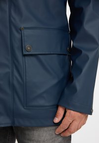 Giacca impermeabile blu marina realizzata in materiale liscio e resistente, con una tasca laterale, dettagli in bottoni argentati e una vestibilità sagomata ai polsi.
