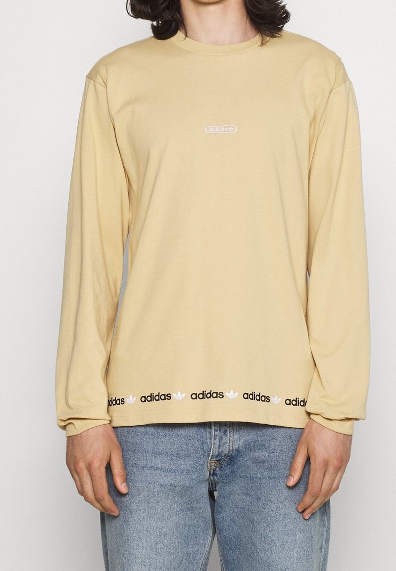 Långärmad beige t-shirt i bomull, med en liten vit Adidas-logga på bröstet och ett svart band med upprepade loggor vid nederkanten.