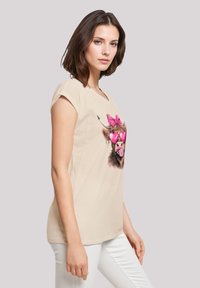 Beige short-sleeve t-shirt featuring a graphic of a cow with pink glasses and bows, paired with white pants; smooth fabric texture.