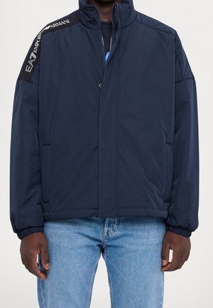 Marineblauwe gewatteerde jas met hoge kraag en zwart schouderstuk met opschrift "EA7 Emporio Armani", gedragen met lichtblauwe spijkerbroek.