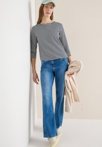 Jeune femme portant une chemise à rayures, un jean bleu, des baskets blanches et une casquette bleu clair, tenant une veste beige, appuyée contre un mur blanc.