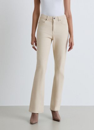 Donna con jeans beige a vita alta e gamba dritta, maglietta bianca infilata e stivaletti marroni con punta appuntita, in piedi davanti a uno sfondo uniforme.