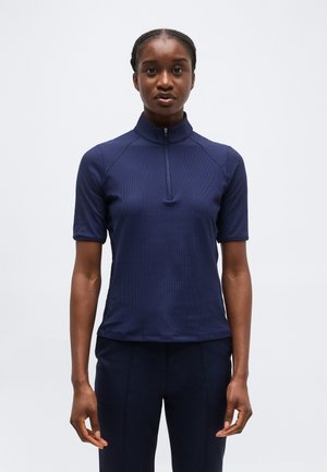 Donna con capelli intrecciati che indossa una maglia blu navy con zip e maniche corte e pantaloni abbinati, in piedi contro uno sfondo chiaro uniforme.